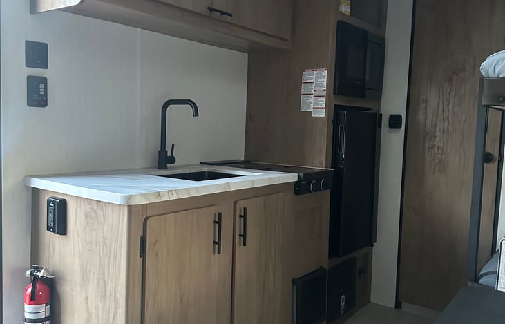 Kitchen preparation area. Sink, stovetop, fridge, freezer, microwave. Also pictured, cabinet storage above and below sink, as a well as a fire extinguisher.