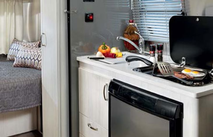 Small and efficient cooking space with sink, 2 burner stove top, microwave, refrigerator with freezer compartment and storage for cookware. Behind is double bed with TV and optional privacy screen.