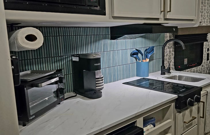 Kitchen area with two stove burners, toaster oven, K cups coffee maker (with K cups), Mini fridge, in counter trash can, sink with hand soap dispenser.