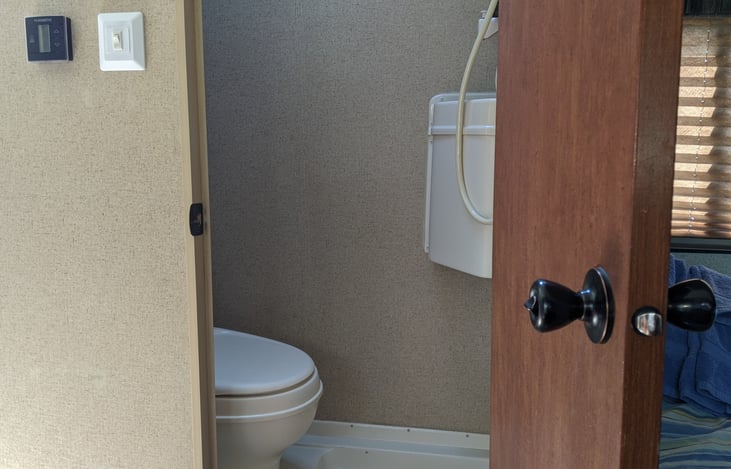 Bathroom with flushing toilet and small sink. Has shower head above sink and electric vent above.