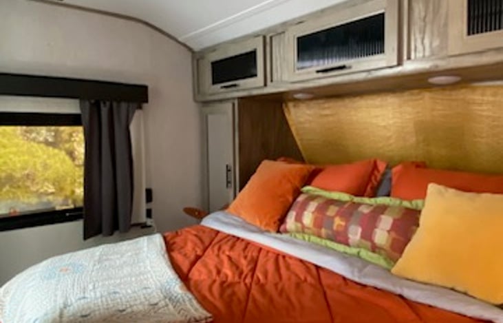 RV Queen bed with overhead storage compartments and emergency egress window.  Recessed lighting above with side- table lamps provided.