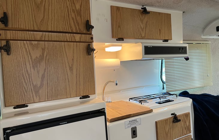Kitchen with propane stove, refrigerator, sink, vent, water pump, cutting board, storage.