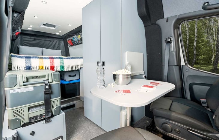 Van Interior with bed folded down. Multiple tables up front and front chairs swivel to enjoy dining or games together.