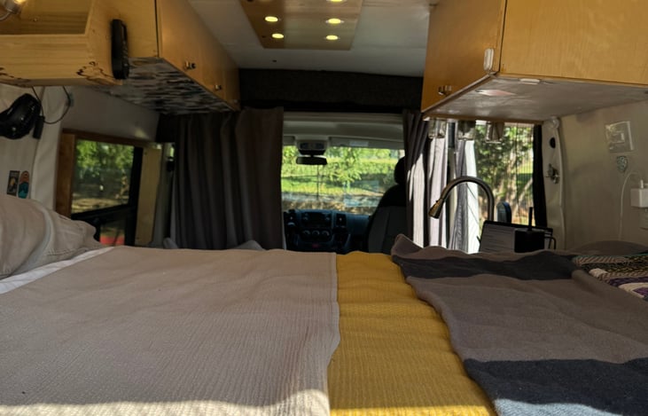Interior view of van from the back facing the cab with bed and upper shelves in view.