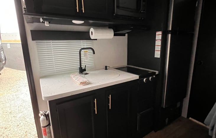 Kitchen has pots/pans/storage containers, coffee mugs, spices, measuring cups, microwave, fridge, two burner gas stove.