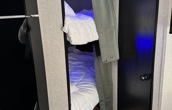 Full-size double bunks featuring luxury gel mattresses and white flannel bedding. Each station includes integrated 120V outlets, adjustable blue or amber reading lights, and privacy curtains.