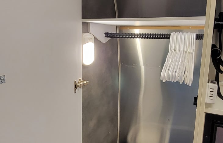 Closet with hangers and a collapsible laundry basket. Additional storage is located over the bed.