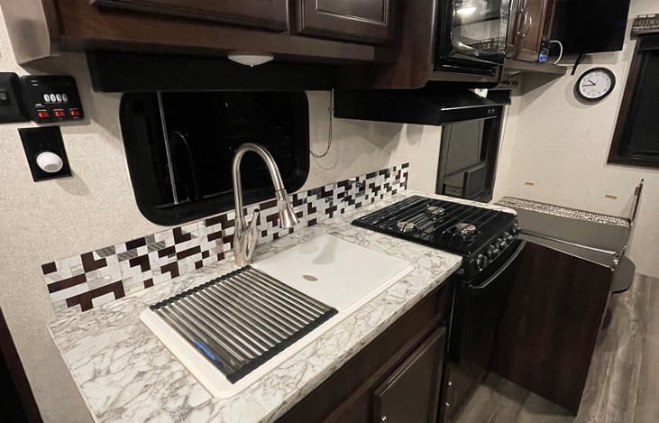 Efficient kitchen with ample storage, gas range, oven, microwave and dual sink with cutting board and dish drying rack.