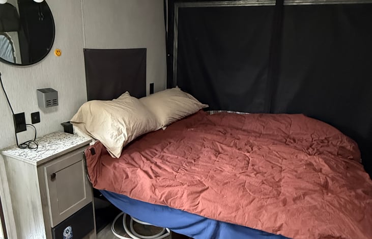 bed side cupboard with mounted mirror and light.  black out shade on window behind top op bed