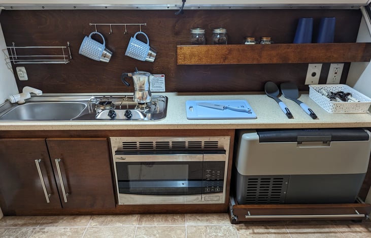 110V Microwave, 12V cooler/freezer, two burner propane stovetop, stainless steel sink, silverware, dishes, pot/pan, coffee maker.