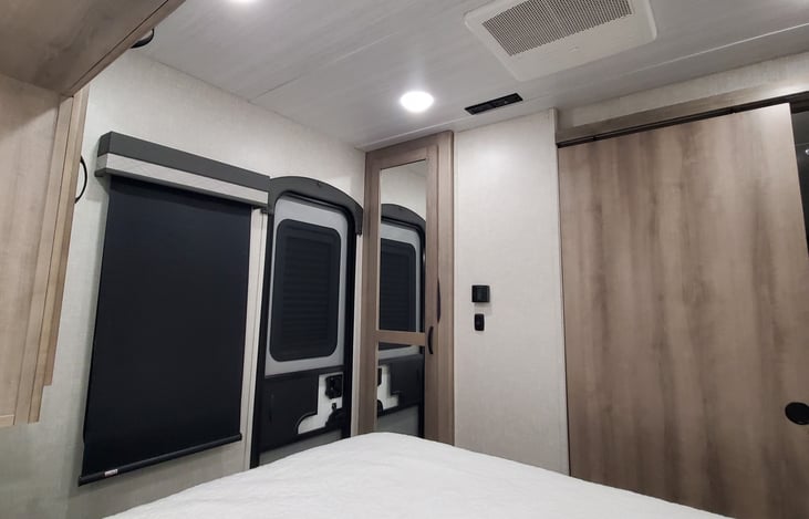 Camper has blackout blinds throughout. The mirrored door is the closet space with an open shoe storage at the bottom. Bedroom has dedicated HVAC. Exit door from bedroom.