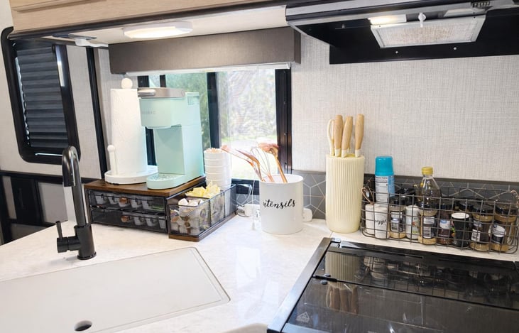 Fully equipped kitchen featuring a deep farmhouse sink with cutting board cover for extra prep space, under-cabinet lighting, three-burner stove, and cooking utensils provided.