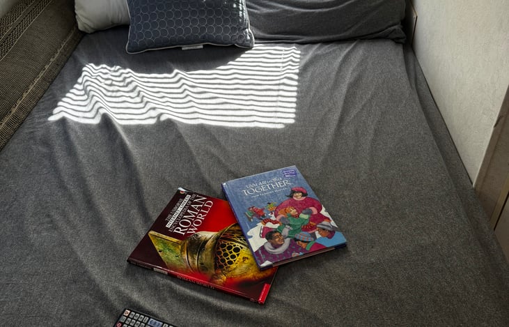 Queen size bed in the back with bedding, pillows, blankets and kids books.