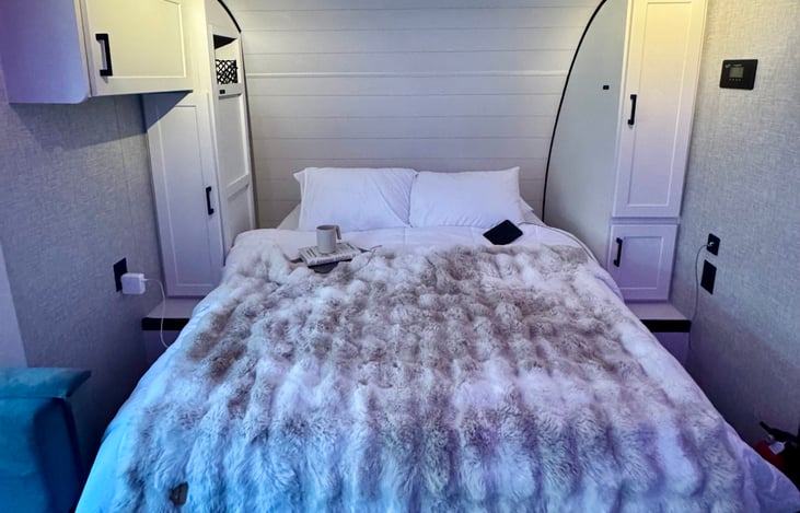 Cozy queen bed featuring crisp white flannel sheets, accompanied by integrated wardrobe cabinets and built-in 120V power outlets.