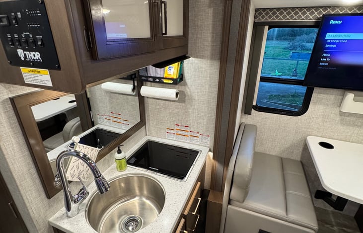 View of sink and stovetop area. Burners have a folding solid cover so that space can be used when burners are not in use. Mounted paper towel holder with storage above.
