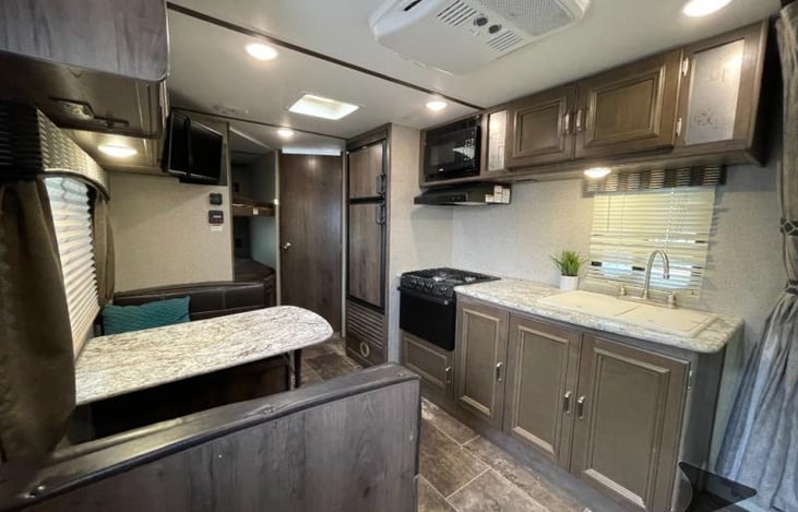Main area of the RV with kitchen with freezer/fridge combo, dual sink, microwave, stove, and oven. Seating booth across from the kitchen area that can seat 4 people and can be converted to a bed.