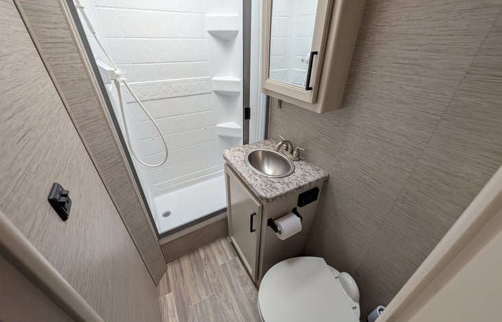 Private rear bathroom features a spacious corner shower with a skylight for extra headroom, a foot-flush toilet, a vanity sink with storage, and a large mirror. Perfectly clean and functional!