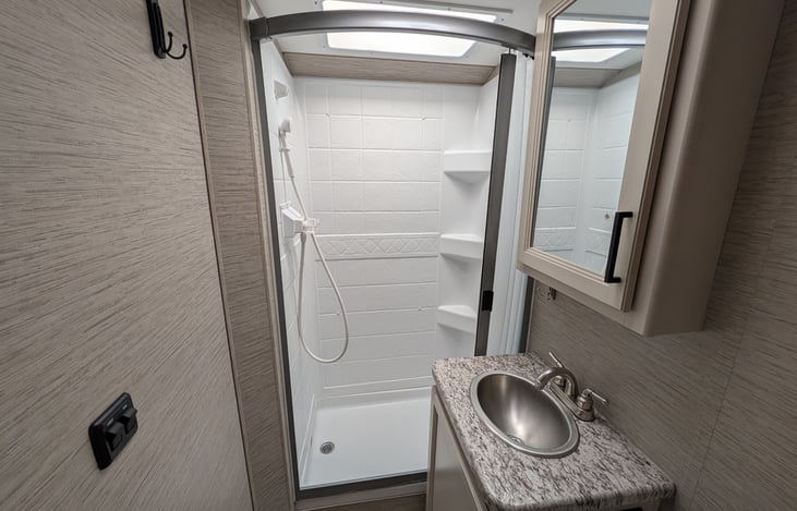 Private rear bathroom features a spacious corner shower with a skylight for extra headroom, a foot-flush toilet, a vanity sink with storage, and a large mirror. Perfectly clean and functional!