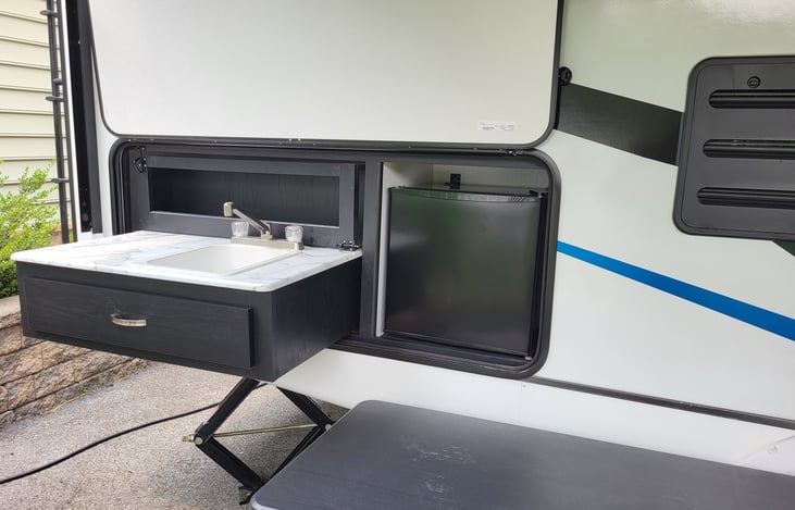 Outdoor kitchen has a pull-out sink with under counter drawer for grilling utensils and a spice rack behind the sink, also a mini refrigerator.