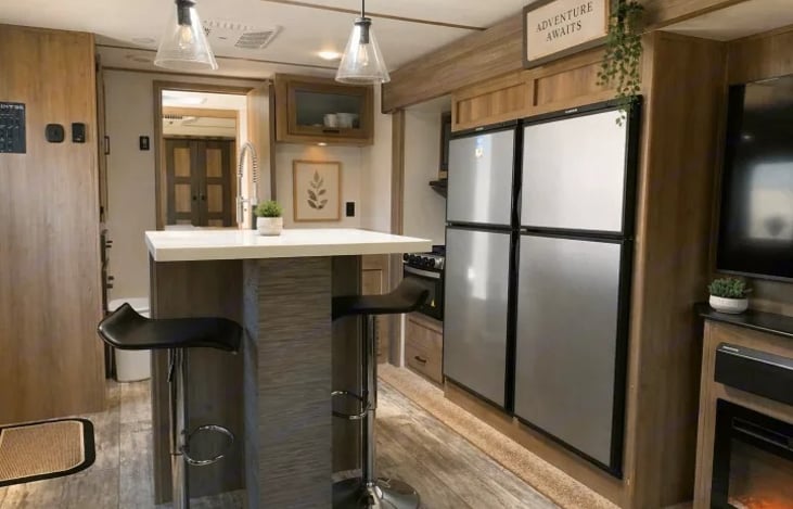 The kitchen is centered around a large island with a stainless steel farm-style sink, a roll-out drying rack, and two bar stools at a raised breakfast bar. It includes a unique dual 7 cu. ft fridge