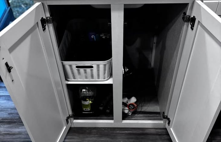 A neatly organized under‑sink cabinet stocked with the essentials you actually need on the road — pots and pans, cooking oil, kitchen towels, a dish‑drying mat, and a cutting board — all arranged for
