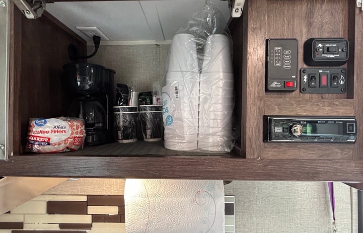 Kitchen upper storage stocked with coffee maker and supplies. To the right is the holding tank level display and pump switch, slide out and control switches, interior radio with Bluetooth capability.