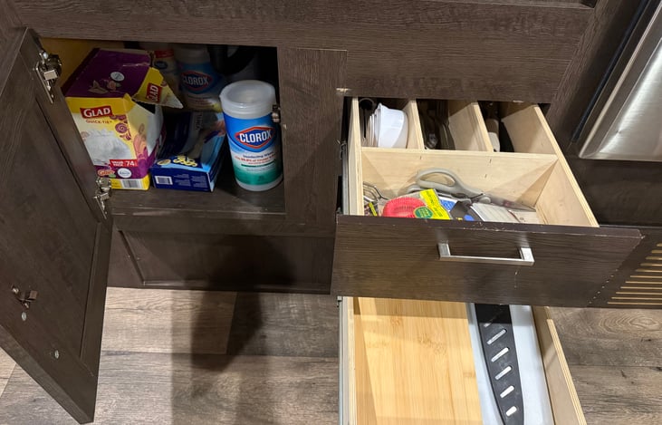 Under sink cabinet contains cleaning supplies and trash bags. Drawers include silverware, measuring cups, scissors, cutting board, and knife