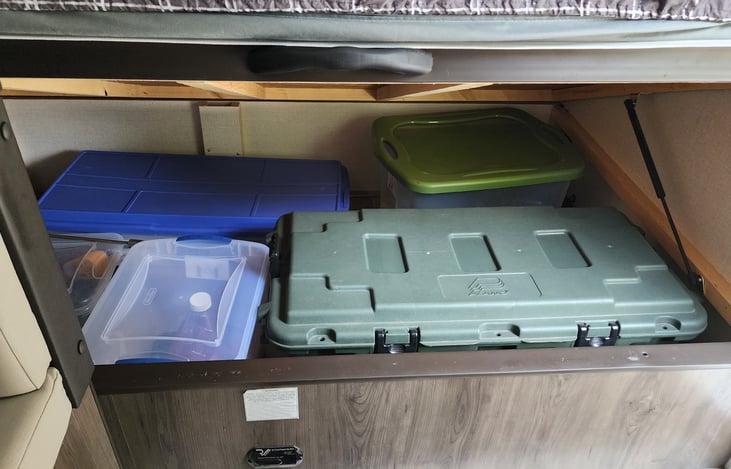 Green tote under bed in storage area has extra pillow and blankets