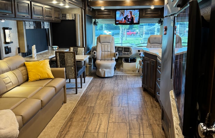 living area from view near bedroom door. There is a sunshade and nightshade at front of rig (both retracted in the image; all living area windows have sunshade and night shades