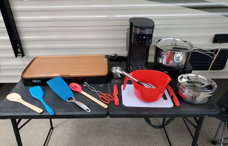 electric griddle, burner, coffee maker (includes refillable k-cup), pots with strainer lid, mixing bowl, whisk, spoon, tongs, flippers, wooden spoon, slotted spoon, cutting board, knives, and table