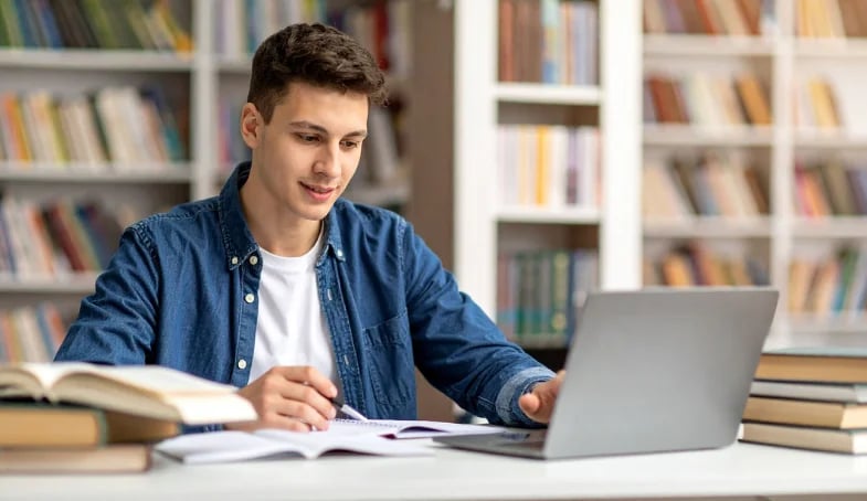 Chico joven sentado en una mesa frente a un ordenador en una biblioteca buscando en todos nuestros libros cristianos pdf