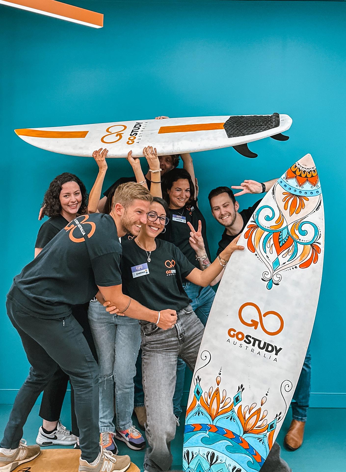 Equipo de Go Study Australia posando con una tabla de surf en su oficina, reflejando la comunidad y cultura australiana de la agencia