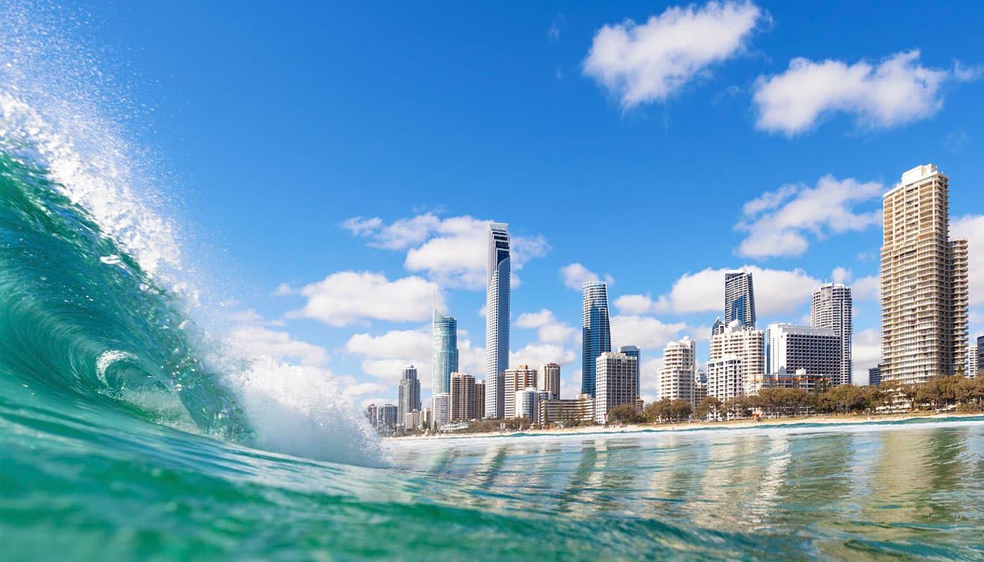 Una ola del océano turquesa rompiendo frente a los rascacielos de la ciudad de Gold Coast, Australia, bajo un cielo azul.
