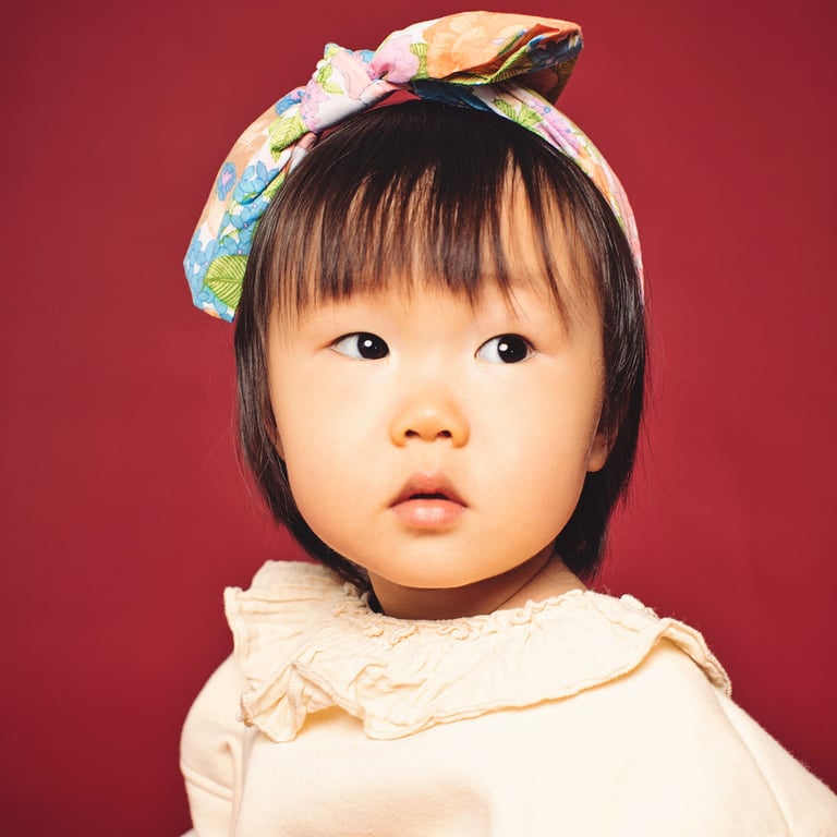 A toddler with a colorful floral bow headband in a cream ruffled top, on a deep red background