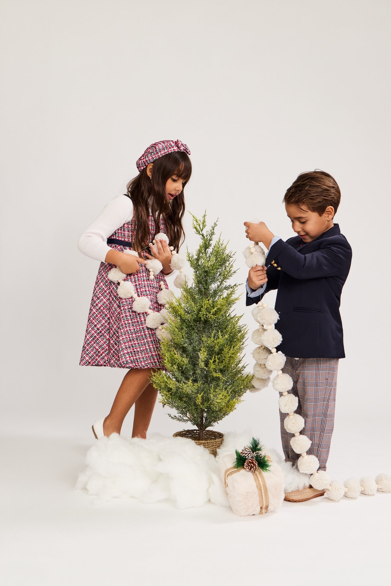 A brother and sister hanging a pom-pom garland on a little tree in fake snow