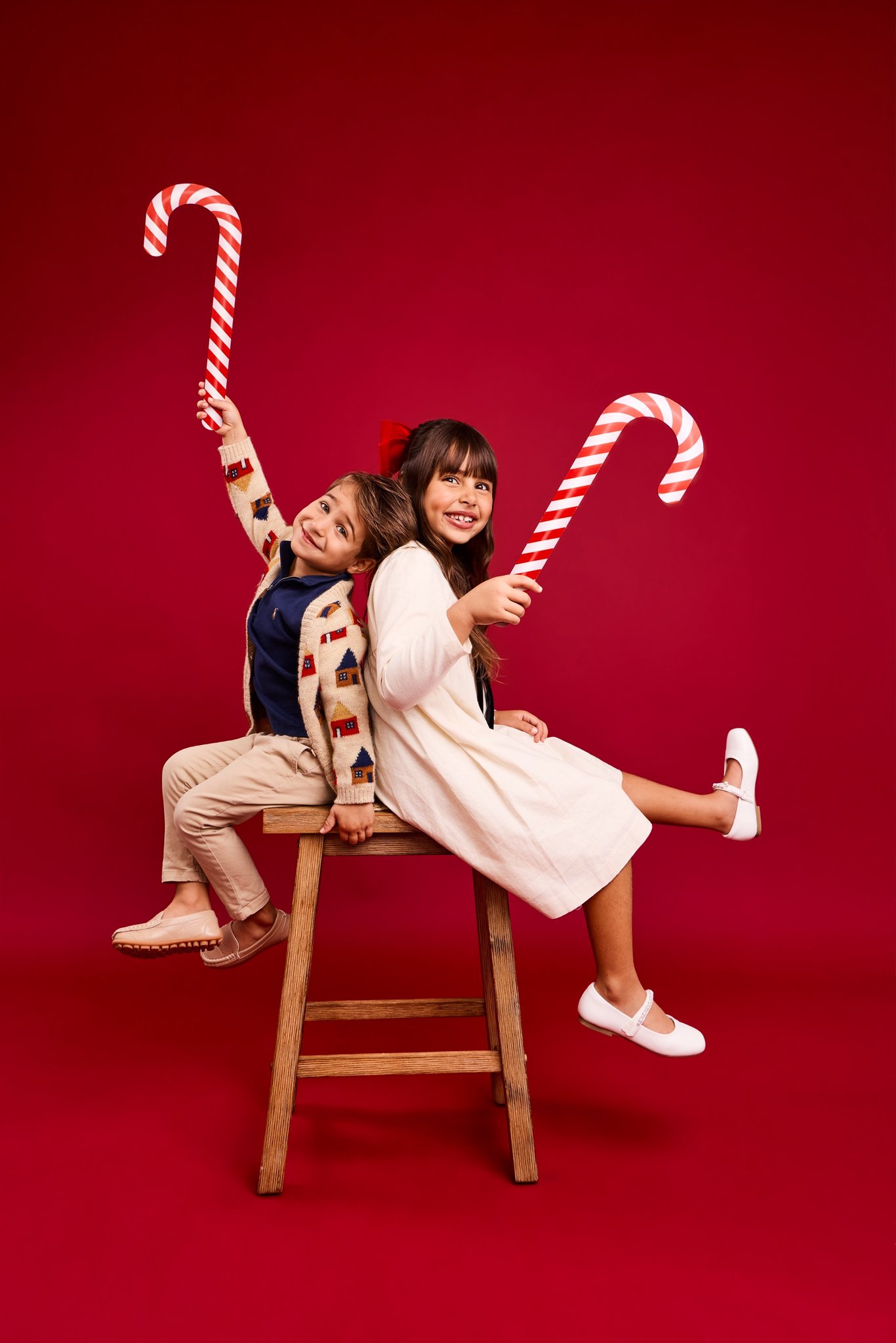 A brother and sister sitting back to back on a wooden stool, waving giant candy canes on a red backdrop