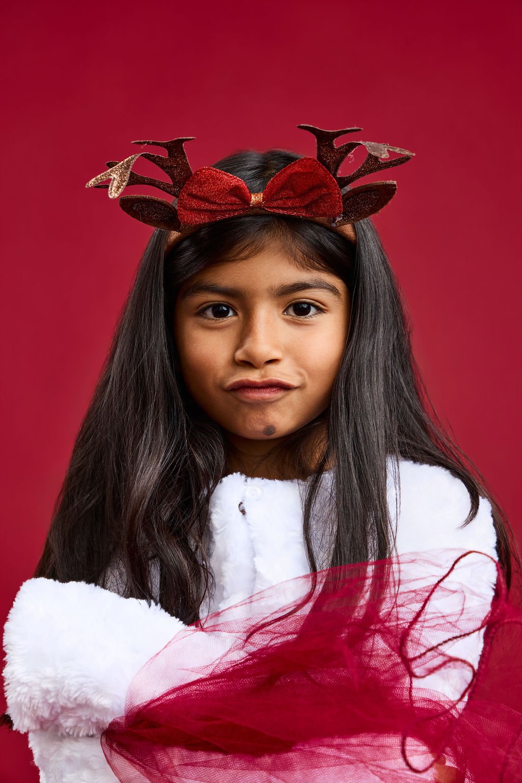 A girl in a sparkly antler headband with a red bow, holding a white faux-fur muff and red tulle, on a red backdrop