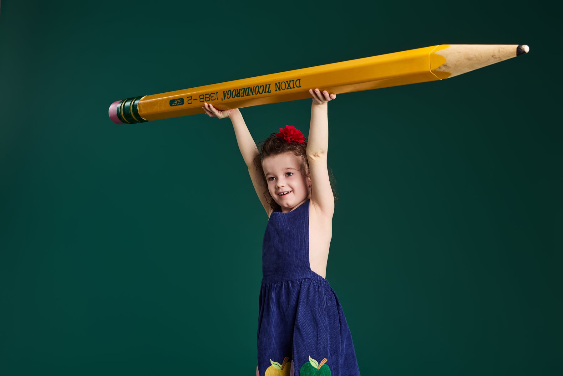 A girl holding a giant pencil overhead on the chalkboard-green set