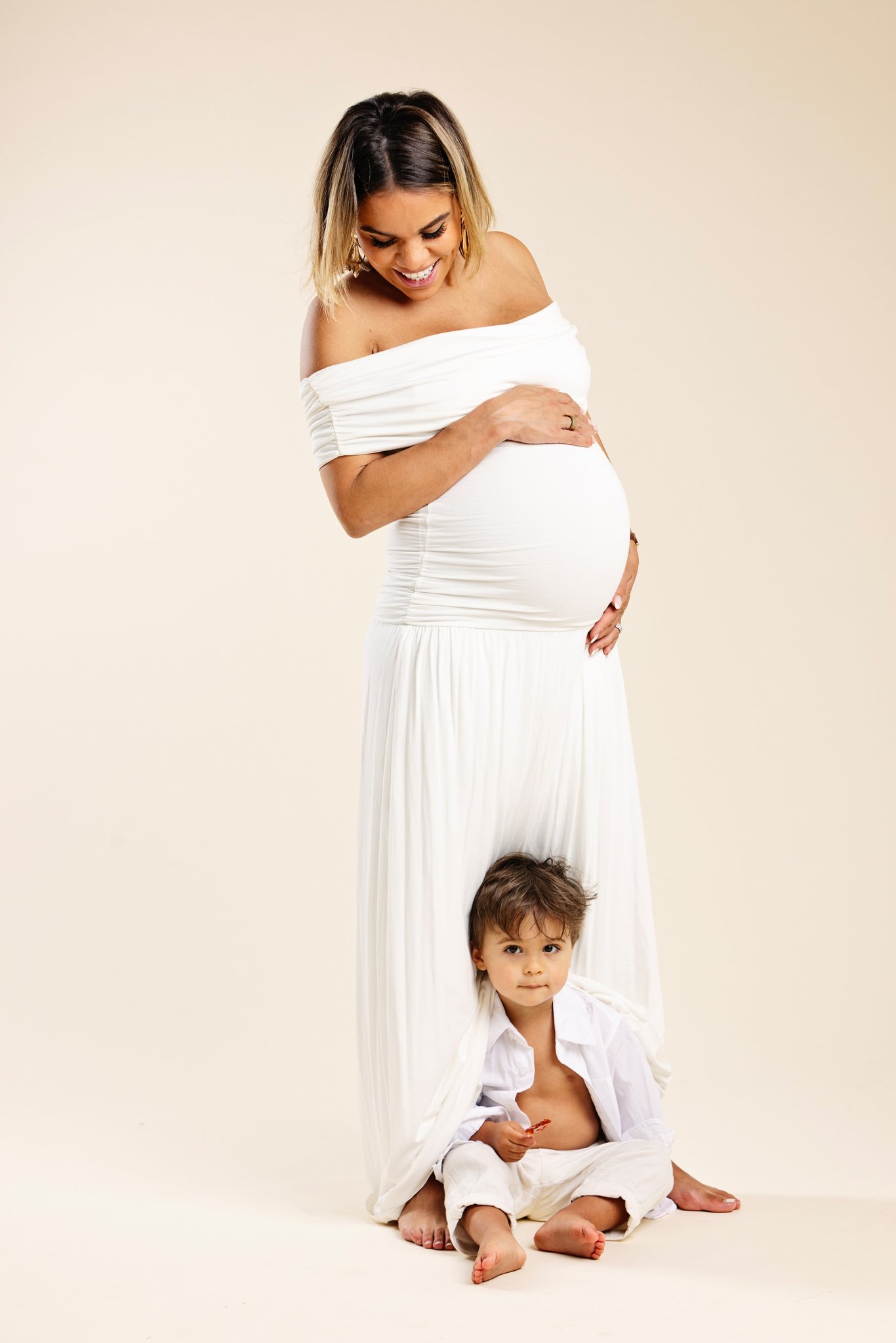An expecting mother in a white dress with her son peeking out beside her