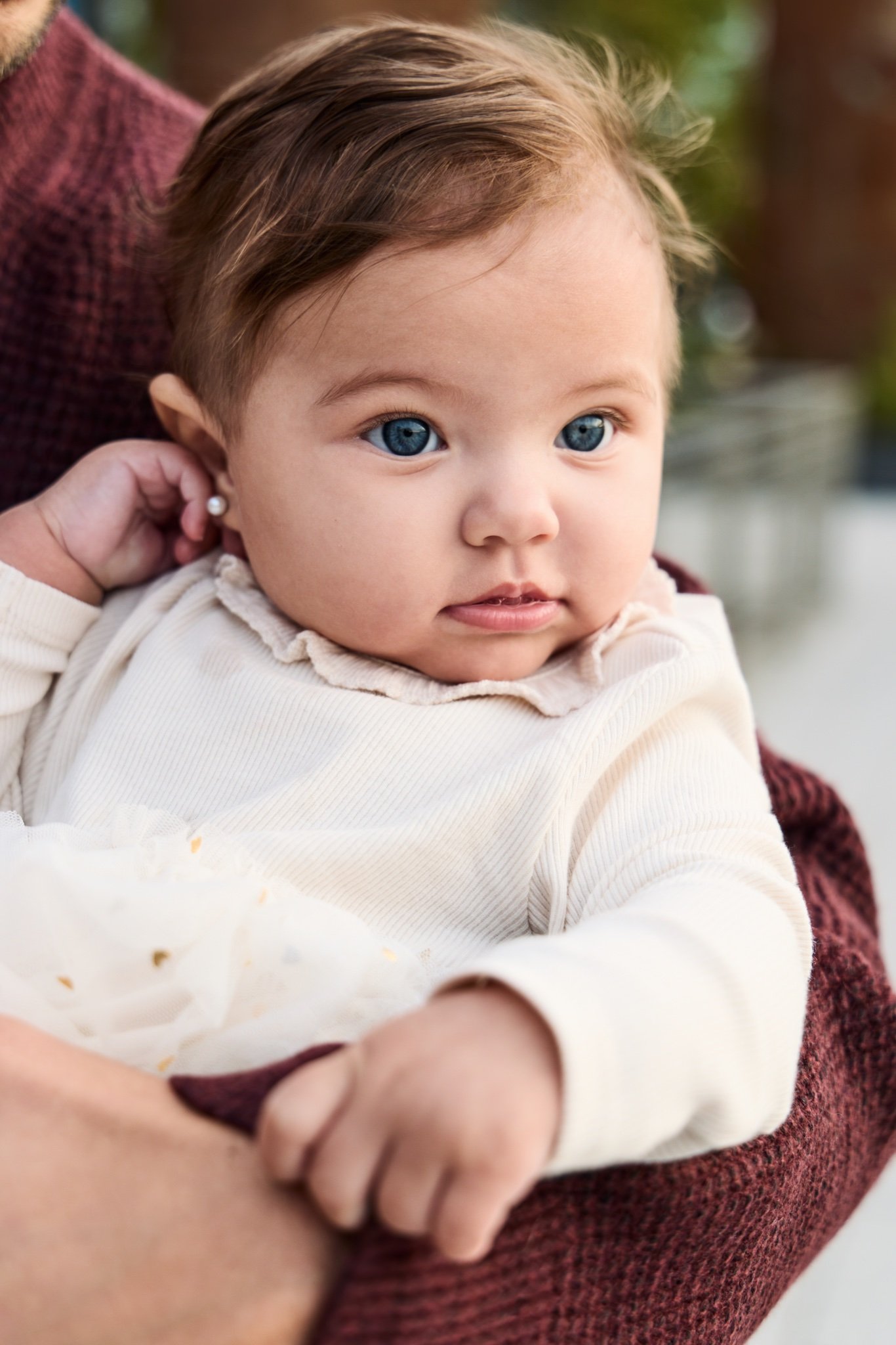 A blue-eyed baby in a cream ruffled collar, held in a parent’s arms