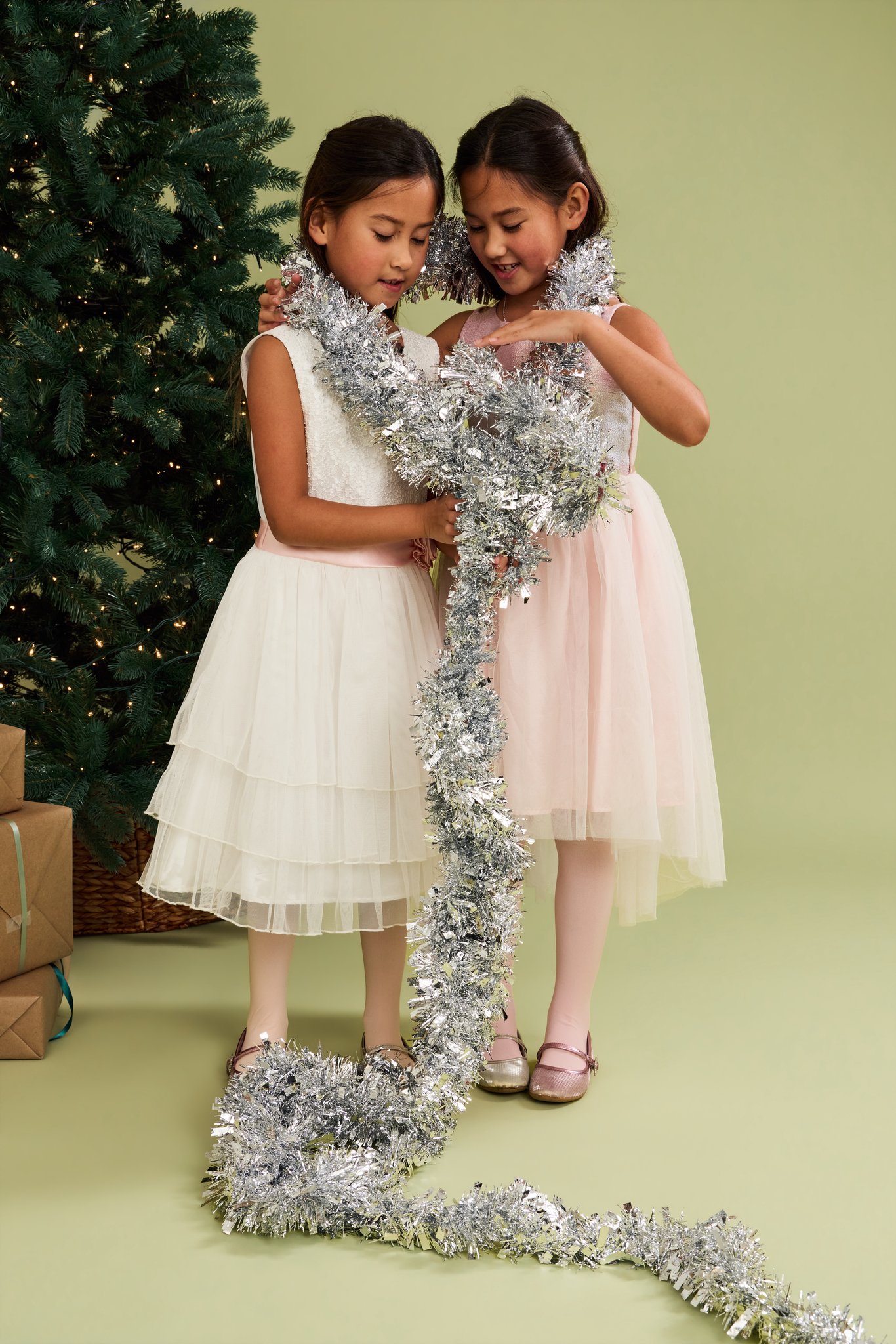 Two girls in white and blush tulle dresses sharing a long strand of silver tinsel beside a lit tree, on the Sage Green backdrop