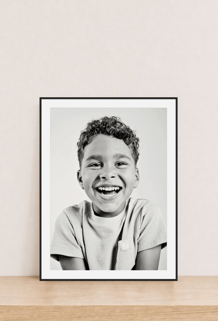 A black gallery frame with a wide white mat holding a black and white portrait of a laughing boy, leaning on a wood shelf