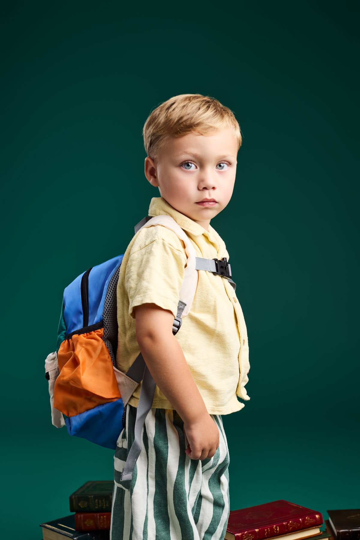 A boy in a yellow shirt and striped trousers wearing a blue backpack, standing beside a stack of books