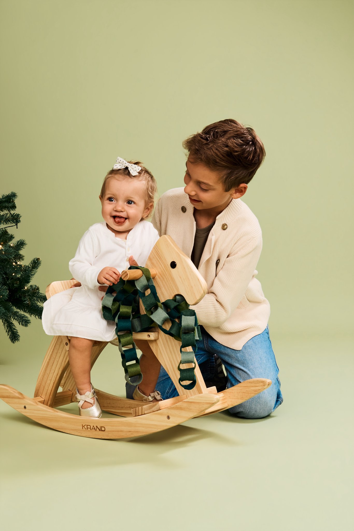 A laughing baby girl on a wooden rocking horse hung with a green paper chain, her brother kneeling beside her, on the Sage Green backdrop