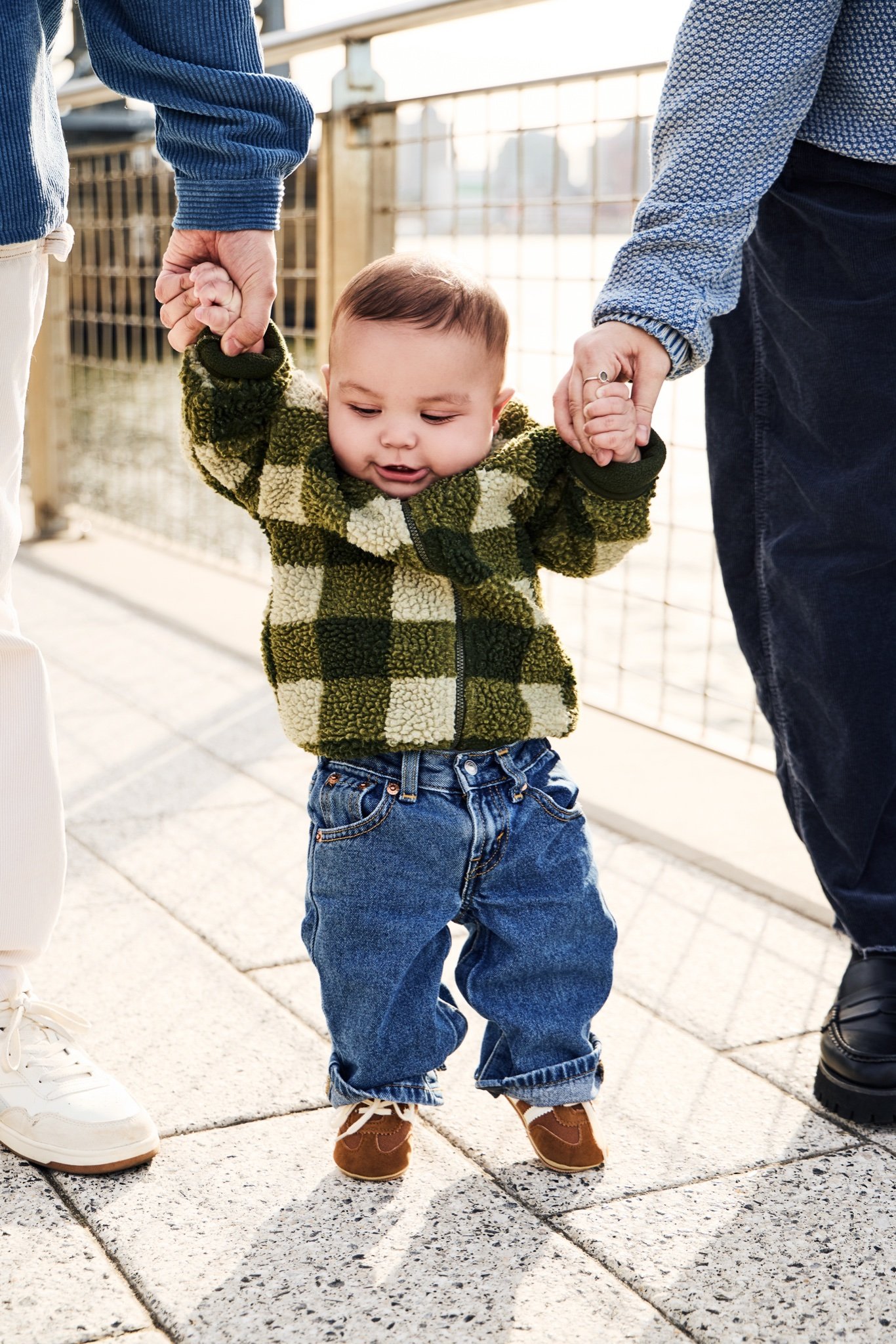 A baby in a green check fleece taking steps along the riverside promenade, holding a parent’s hand on each side