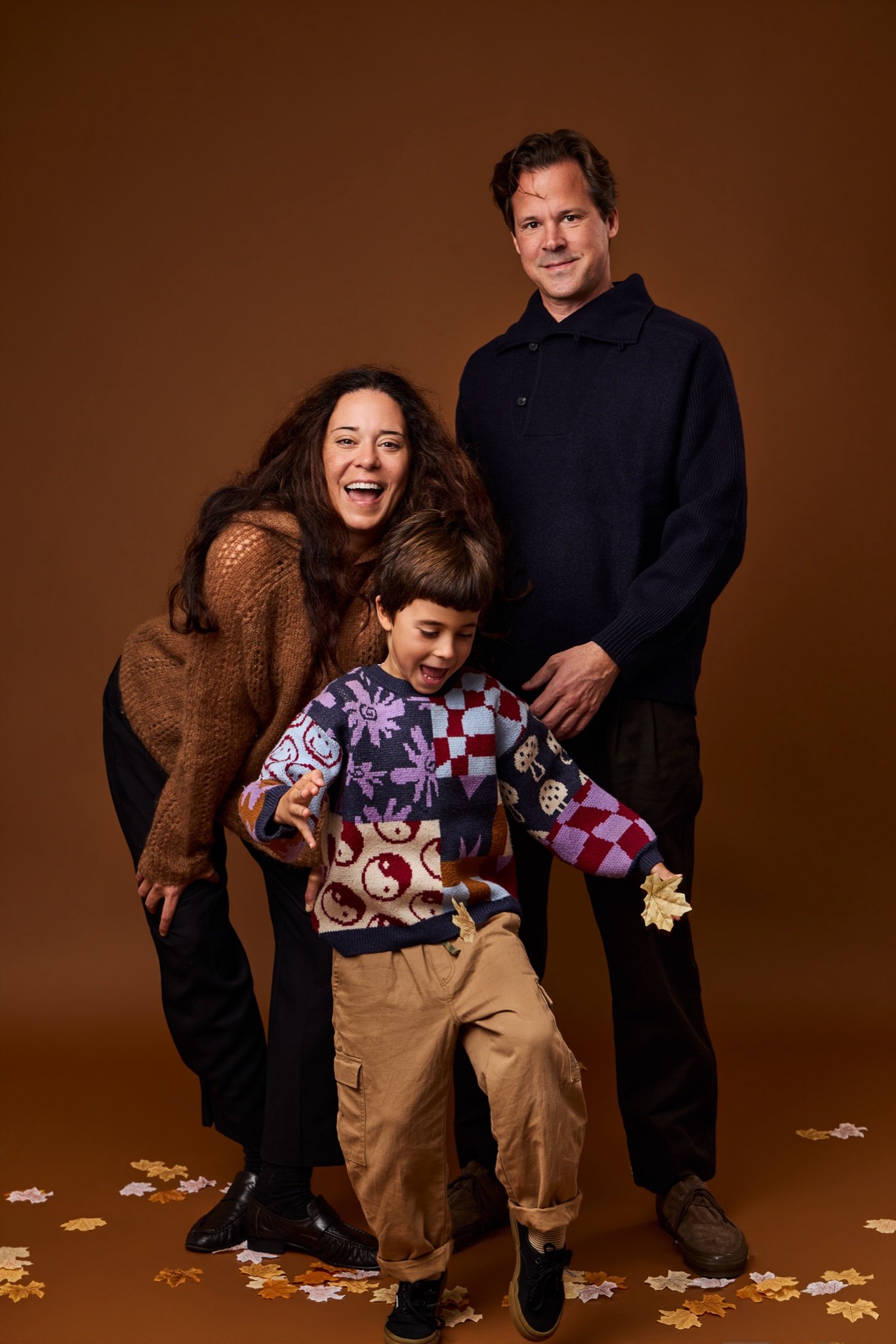 A mother and father laughing behind their son as he steps forward in a patterned sweater, holding a fall leaf, on the Cinnamon backdrop