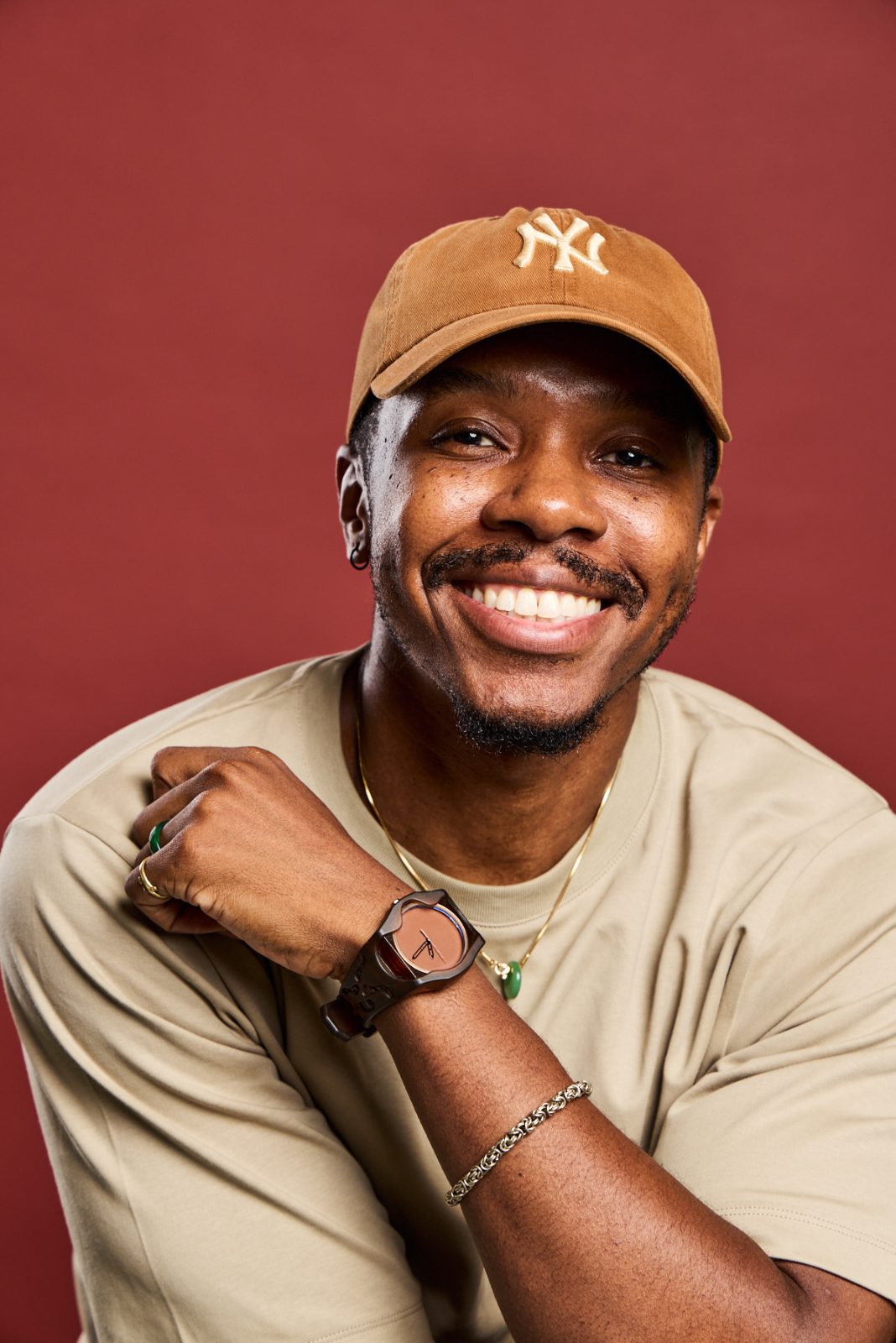 Headshot of a smiling man in a tan cap and t-shirt on a deep red backdrop