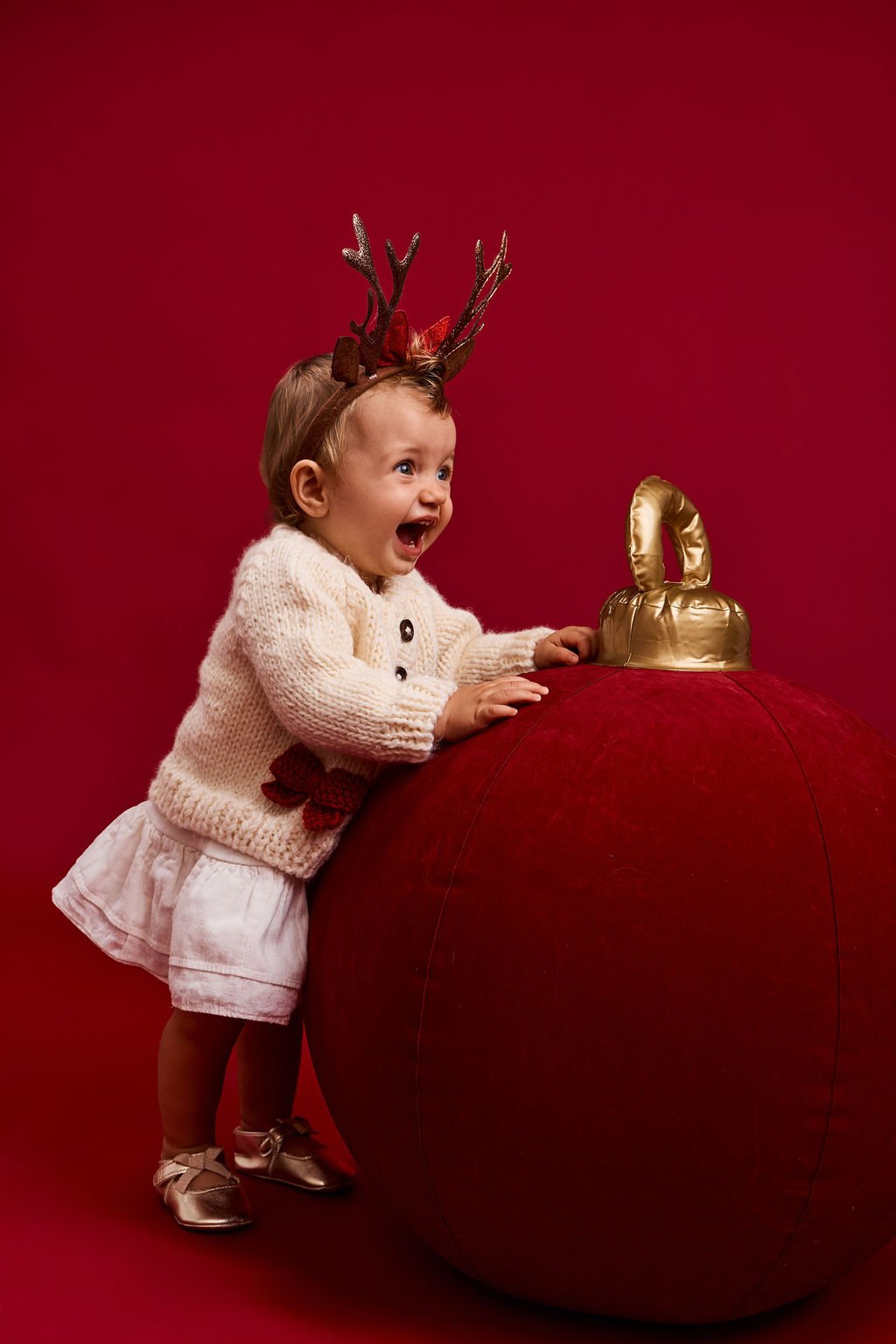 A baby in a reindeer antler headband and cream cardigan gasping beside a giant red velvet ornament, on a red backdrop