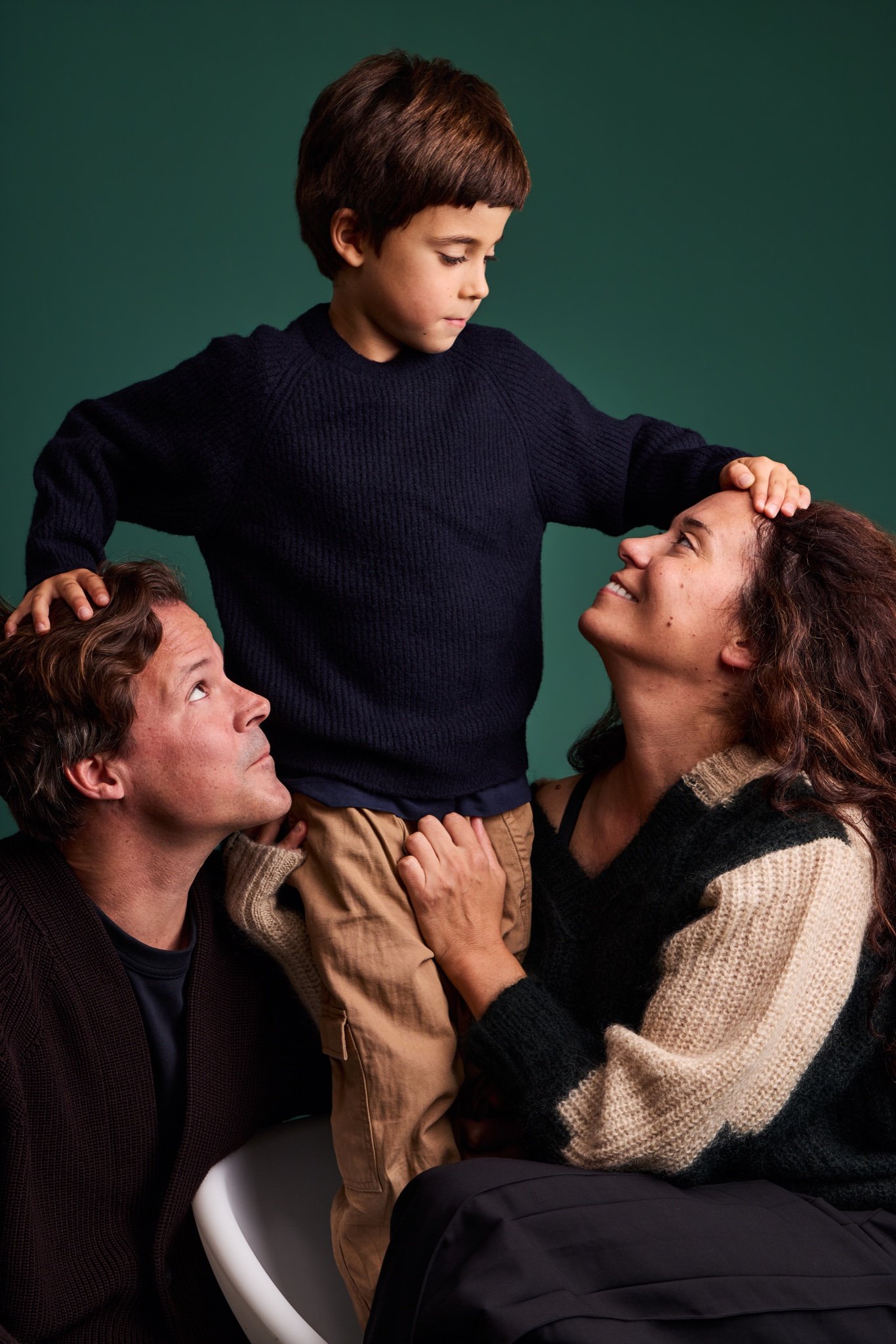 A boy standing between his parents with his hands on their heads as they look up at him, on the Evergreen backdrop