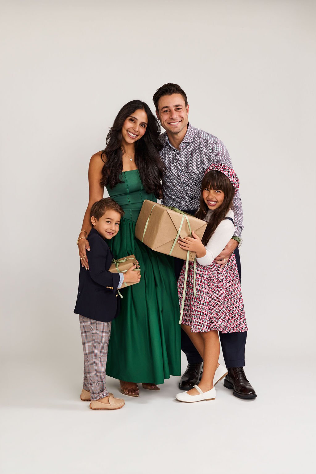 A family of four smiling together on a white backdrop, the mother in a green gown and the daughter holding a wrapped gift